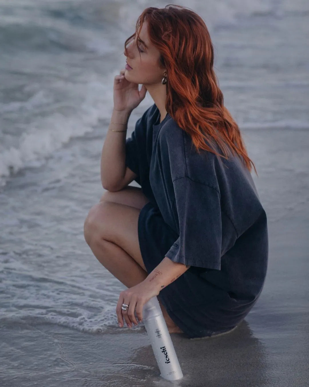 Frau kniet am Einstieg zum Meer und stützt sich dabei auf einer silbernen Haarspray-Dose ab