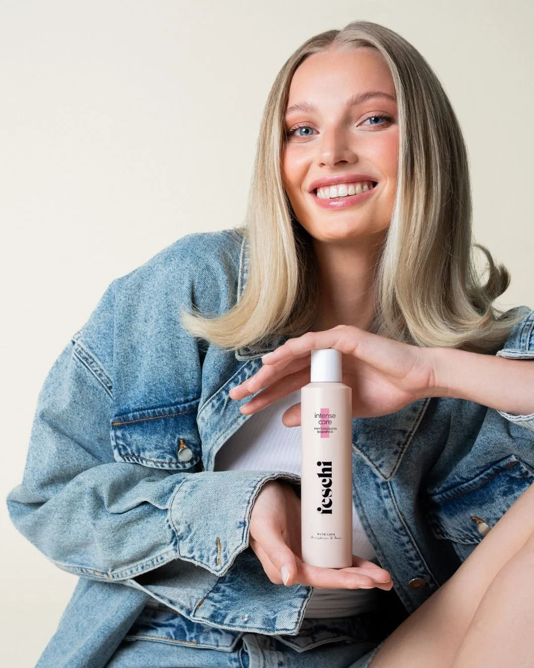 Blonde Frau lĂ€chelt in Kamera und hĂ€lt ein feschi Shampoo fĂŒr strapaziertes Haar in der Hand