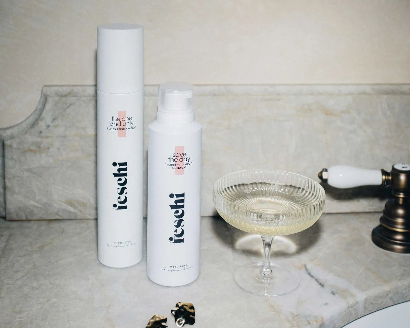 Zwei Trockenshampoos von feschi stehen neben Waschbecken und Champagnerglas