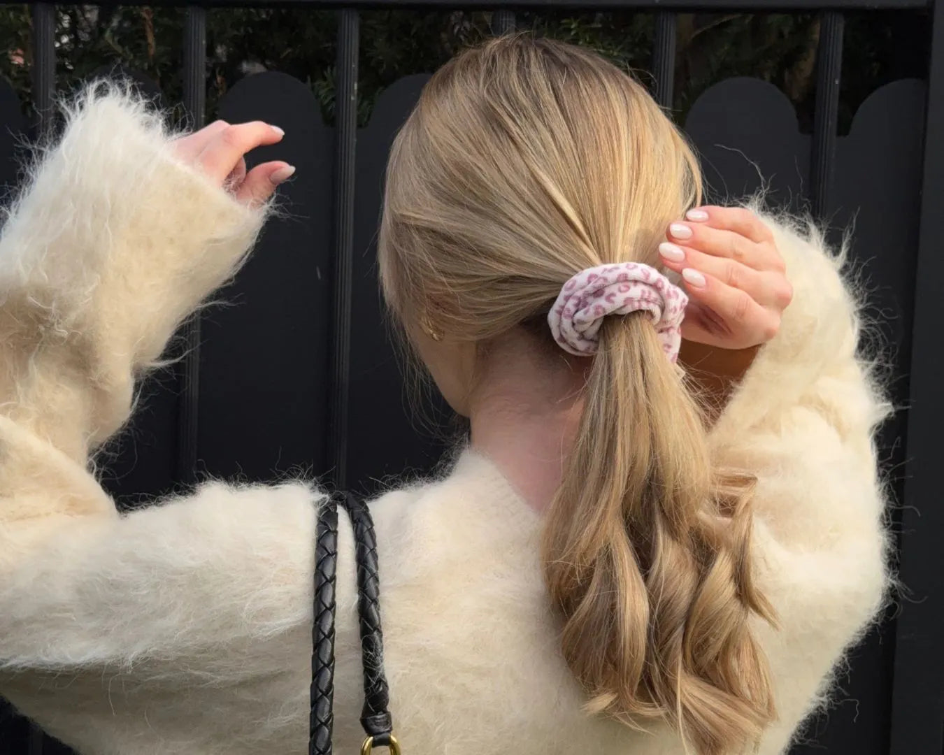 Frau vor schwarzem Zaun mit beigem Wollpulli greift Richtung Kopf und sie hat einen gelockten Zopf mit einem Leo Scrunchie von feschi