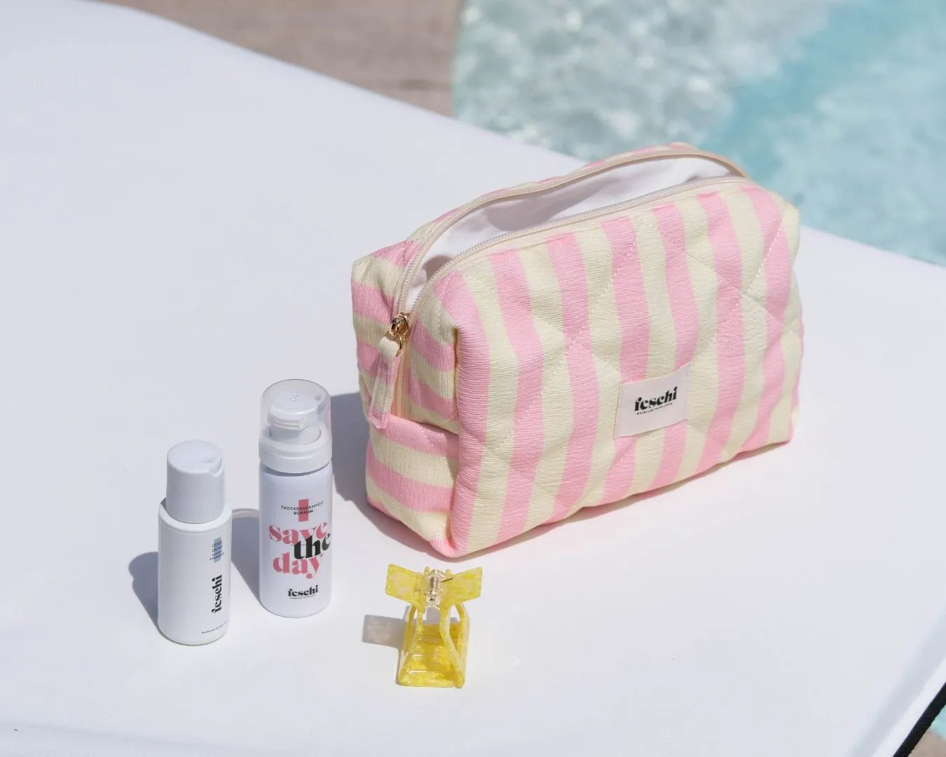 Gestreifte Kosmetiktasche steht auf Sonnenliege und zwei feschi Produkte und gelbe Haarspange liegen daneben