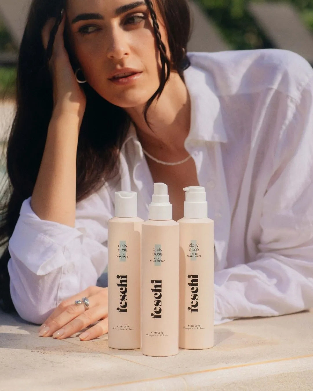 dunkelhaarige Frau liegt beim Pool und schaut aus dem Bild raus. Vor ihre stehen das daily dose Shampoo, Conditioner und Pflegespray von feschi. Die Produkte sind im Fokus