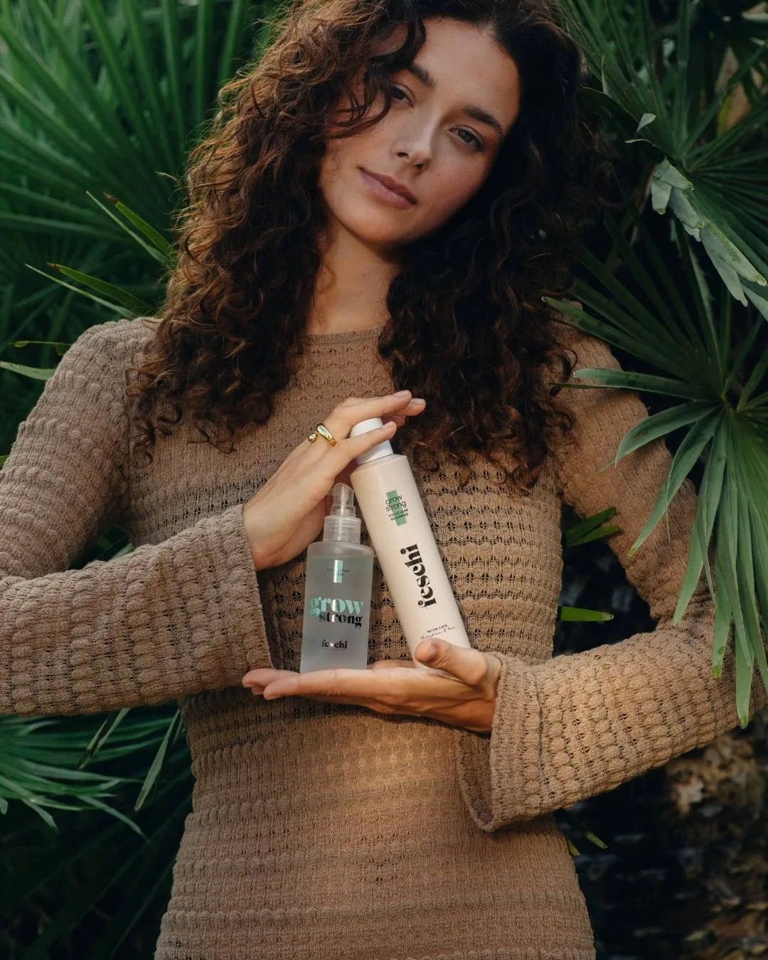 braunhaarige lockige Frau steht vor grüner Pflanze und präsentiert das grow strong Shampoo und Tonic von feschi in ihren Händen