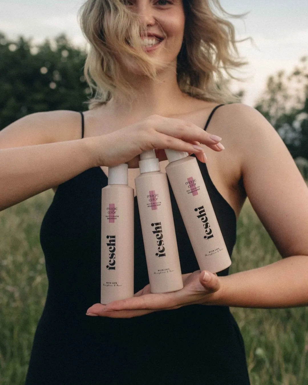 blonde, lÀchelnde Frau mit welligem Haar steht auf einer Sommerwiese und prÀsentiert das magic color Shampoo, Conditioner und Plegespray von feschi in ihren HÀnden.