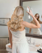Frau steht in Badezimmer und sprüht ihre Haare mit Anti Frizz Spray von feschi an