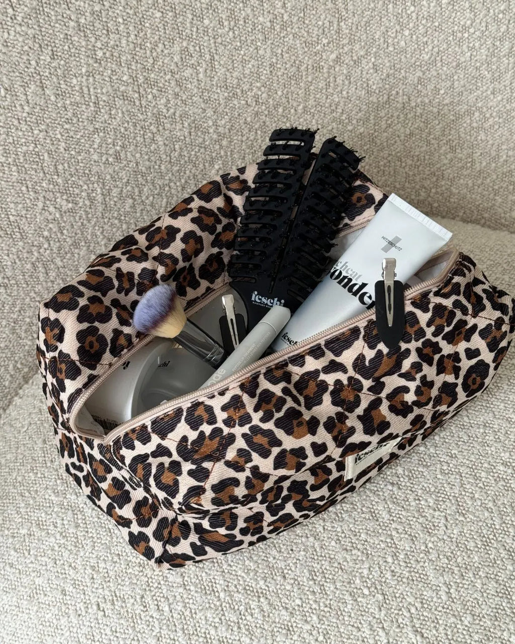 Leopard Beauty Bag von feschi steht auf beigem Polster-Möbelstück und ist prall gefüllt mit Beauty Produkten von feschi und Co. 