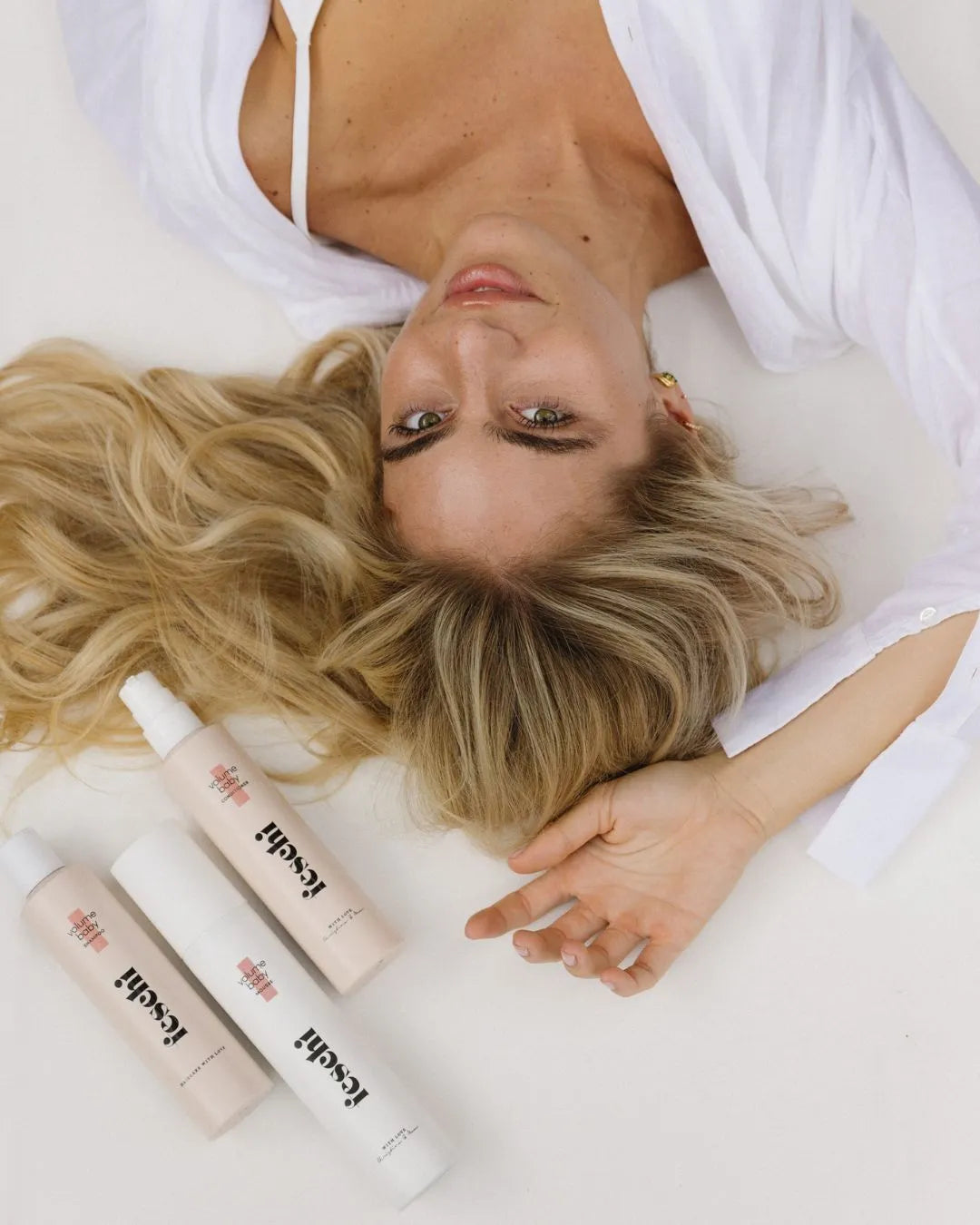 Frau mit blonden langen Haaren liegt auf weißer Unterlage und die feschi Volumen Produkte liegen über ihr