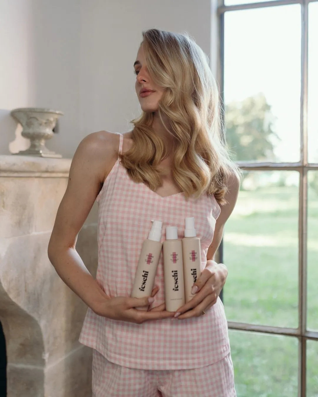 Blonde Frau mit Locken steht in rosa Pyjama und hält drei feschi Produkte in der Hand