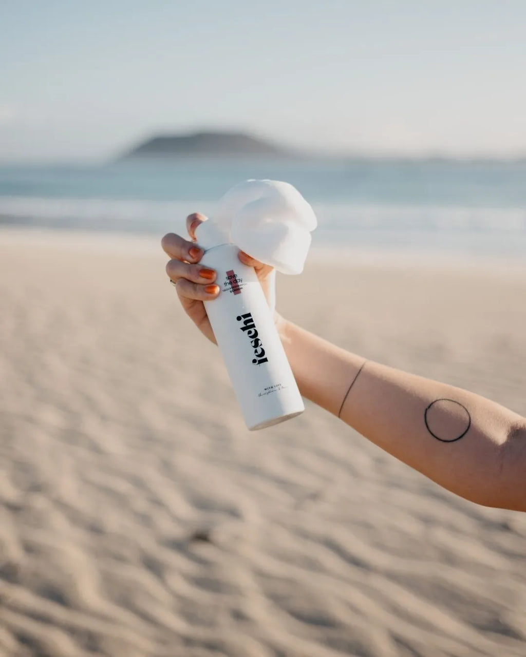 Hand hält das feschi Save the Day Trockenshampoo-Schaum und drückt den Schaum heraus, im Hintergrund Strand und Meer