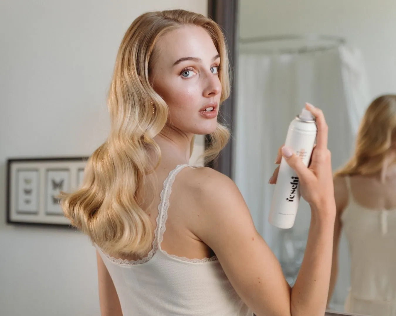 Frau mit langen blonden, welligen Haaren von hinten, blickt nach vorne in die Kamera und hält das feschi Smooth & Shine Anti-Frizz Spray in der Hand