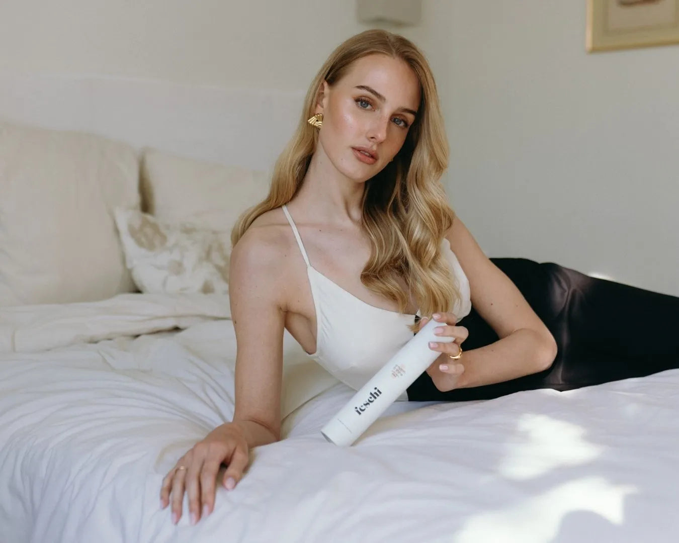 Blonde Frau lehnt seitlich im Bett und hält feschi Anti Frizz Spray in der Hand