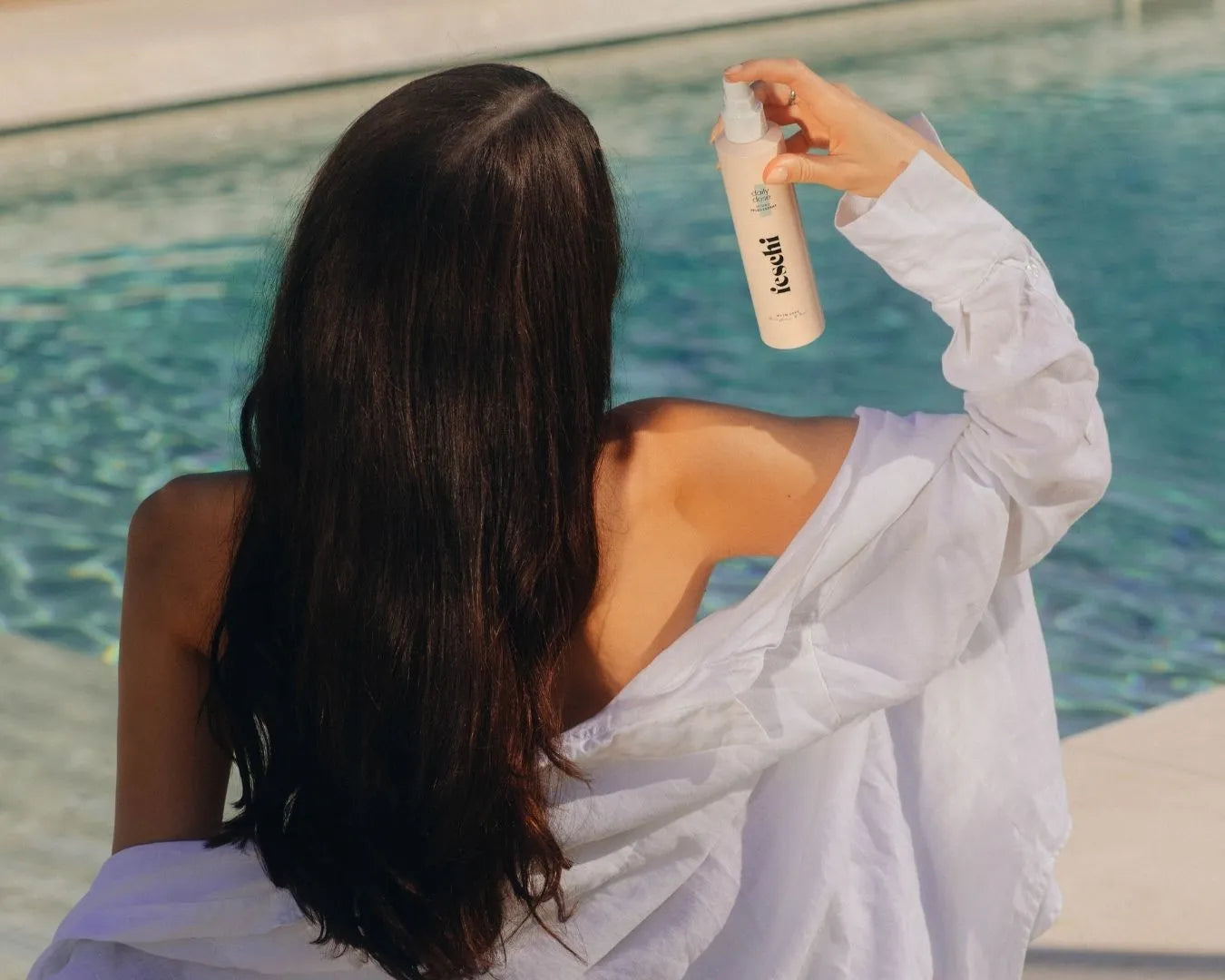 Person mit langen, dunklen Haaren sitzt auf Poolrand und hält das feschi daily dose - Hydro Pflegespray schräg neben ihrem Kopf
