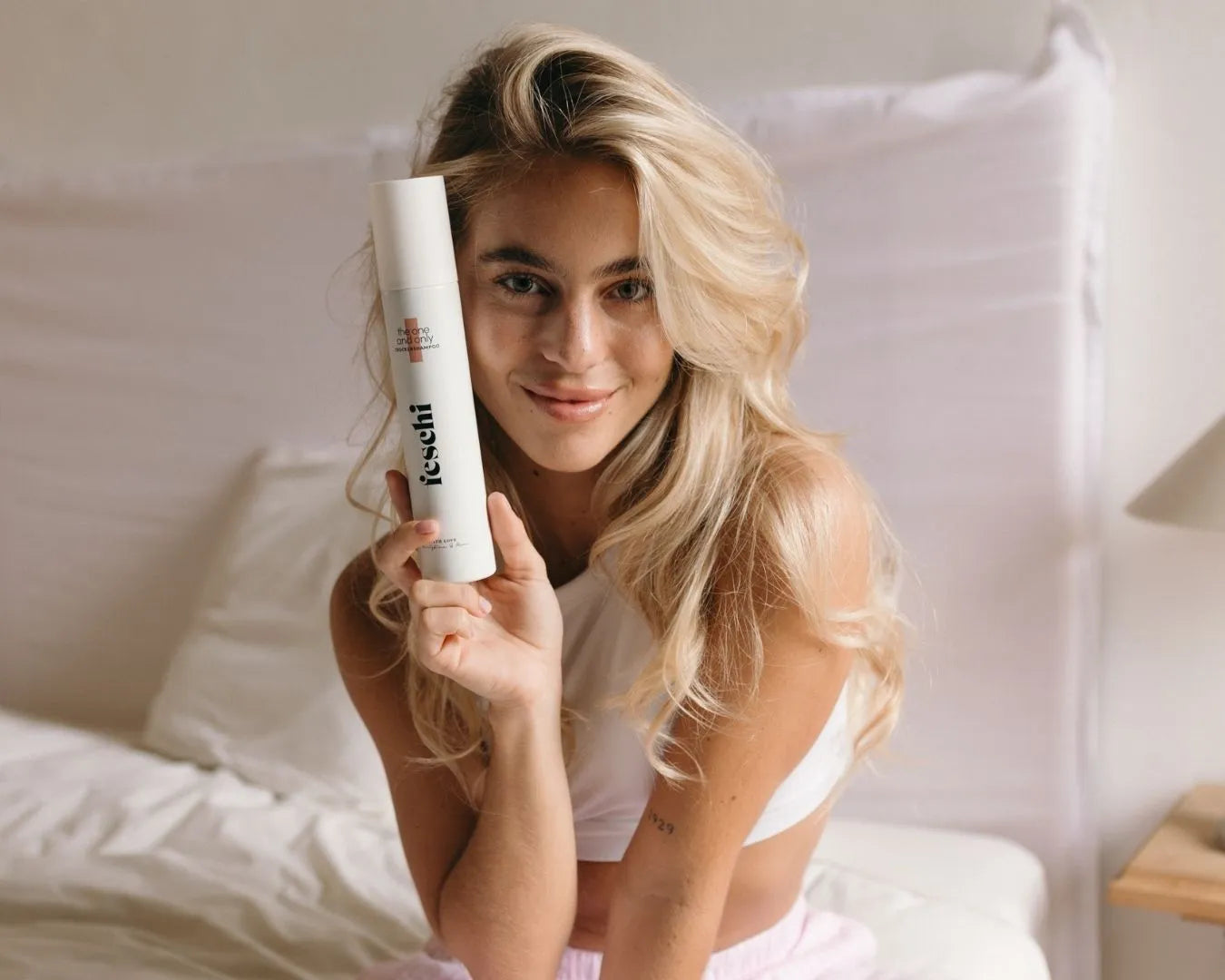 Frau mit langen, blonden Haaren sitzt auf Bett und hält das feschi Trockenshampoo Spray in der Hand