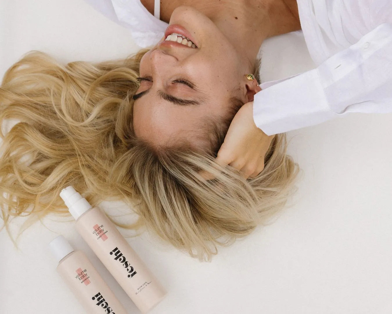 Frau mit blonden Haaren liegt im Bett, neben ihrem Kopf liegen die feschi Volume Baby Produkte: Shampoo und Conditioner