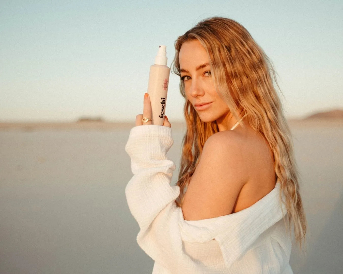 Frau mit langen blonden Beach Waves, stehend am Strand bei Sonnenuntergang, hält das feschi Intense Care Repair Spray in der Hand