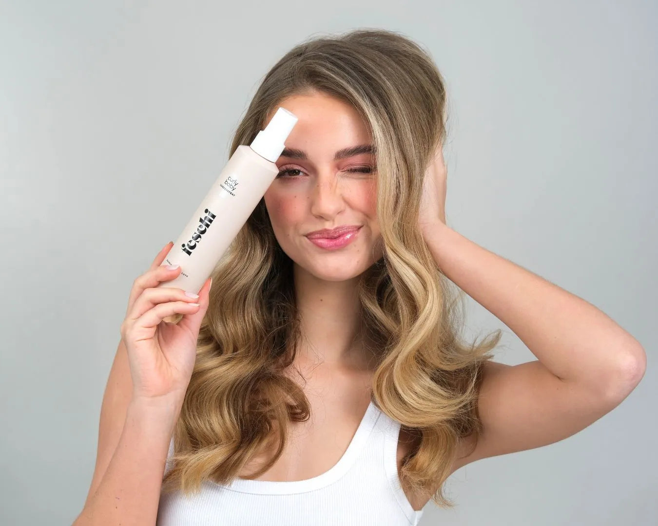 Frau mit blonden, lockigen Haaren hält curly baby Lockenspray in der Hand und zwinkert. 