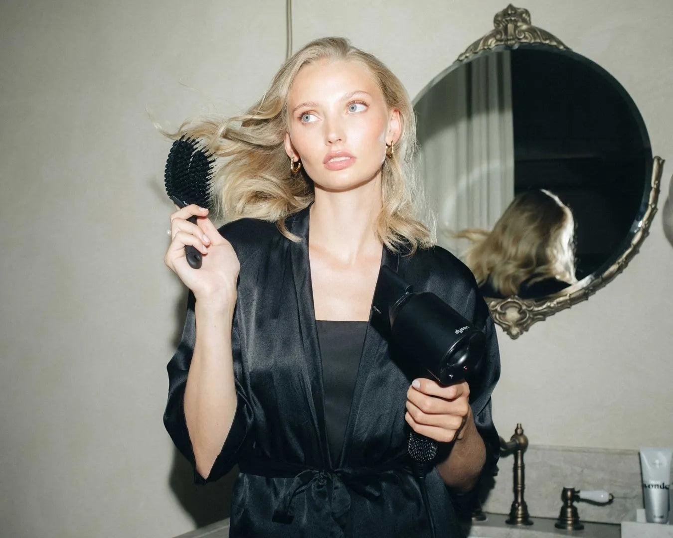 Frau mit langen blonden Haaren hält in der einen Hand einen Föhn, in der anderen die feschi The Essential Hair Brush und blickt zur Seite