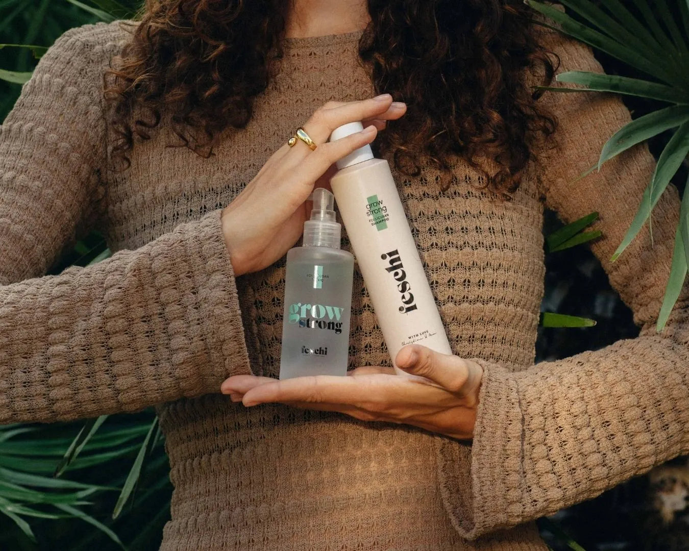 Frau in braunem Kleid vor Palmen hält überkreuzt vor der Brust die Produkte Grow Strong Tonic und Grow Strong Shampoo feschi, Kopf nicht im Bild.