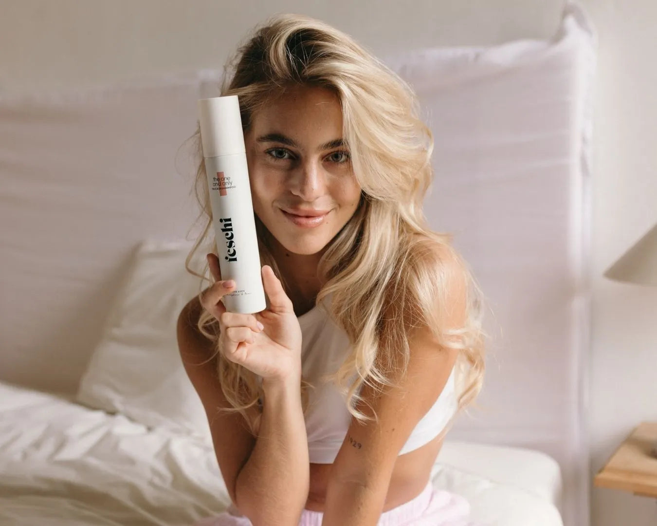 Frau mit langen blonden, welligen Haaren sitzt im Bett und hält den feschi The One and Only Trockenshampoo-Spray in der Hand