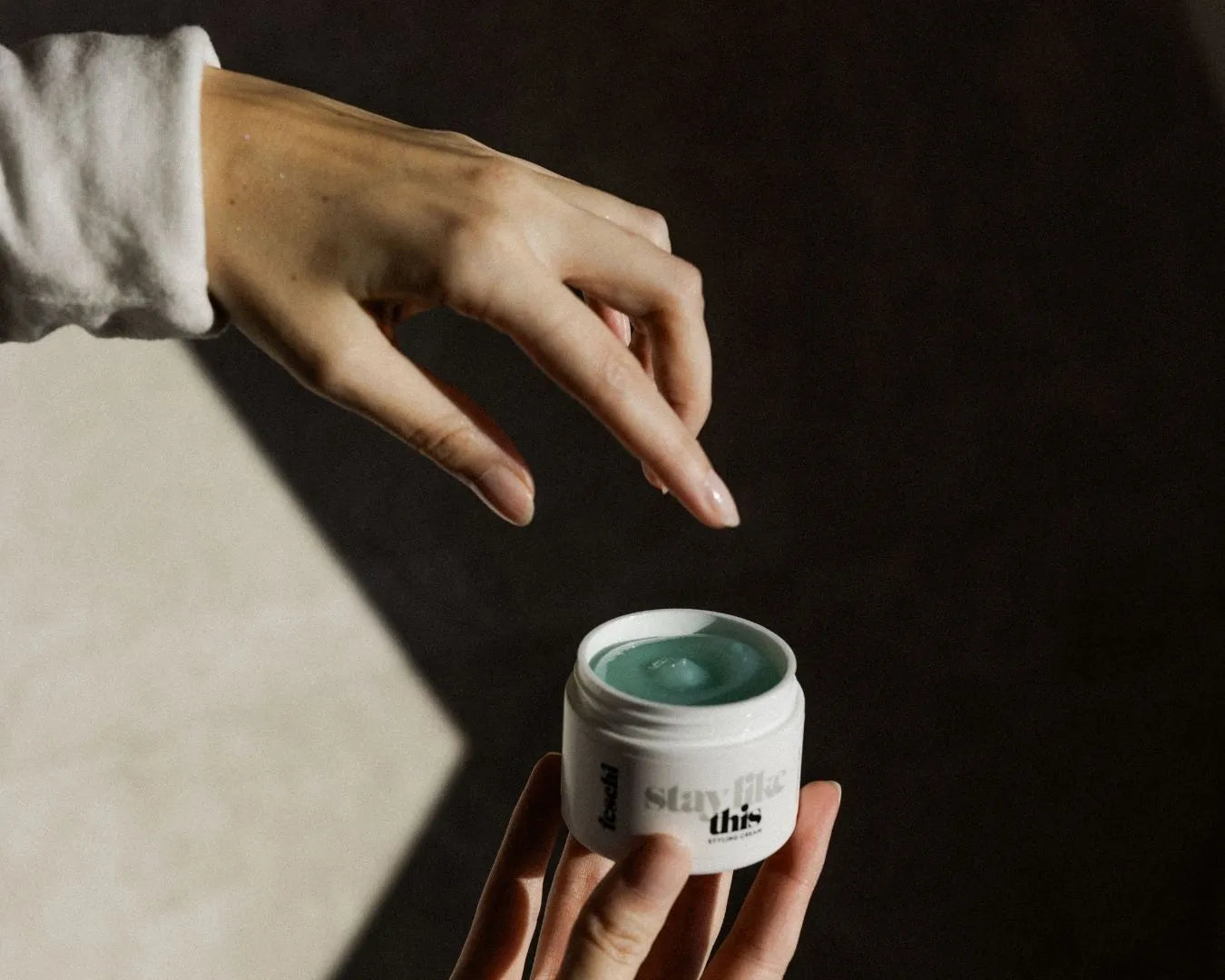 Zwei Hände, eine hält die feschi Stay Like This Styling Cream, die andere greift nach dem offenen Produkt