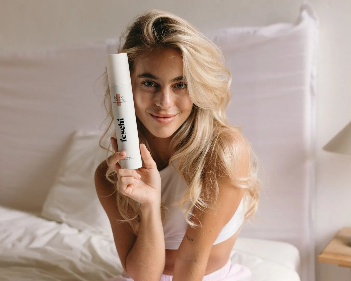 Frau mit langen blonden, welligen Haaren sitzt im Bett und hält den feschi The One and Only Trockenshampoo-Spray in der Hand