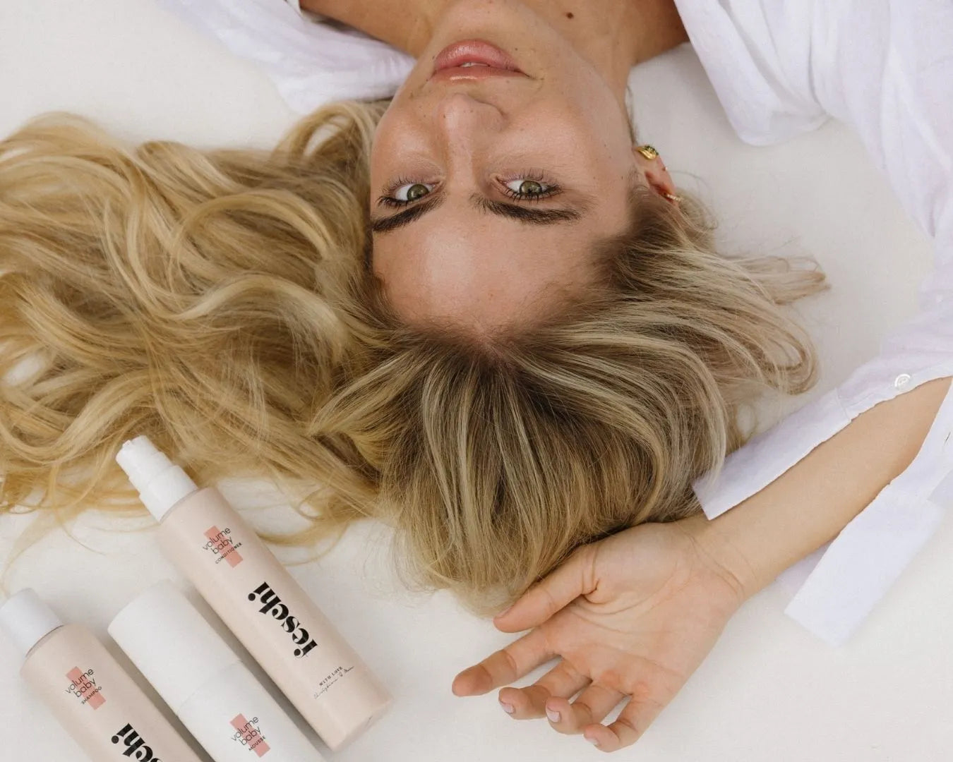 Frau mit blonden Haaren liegt auf weißer Oberfläche, ihre Haare sind sehr schön aufgelegt und über ihr liegen drei feschi Produkte für Volumen