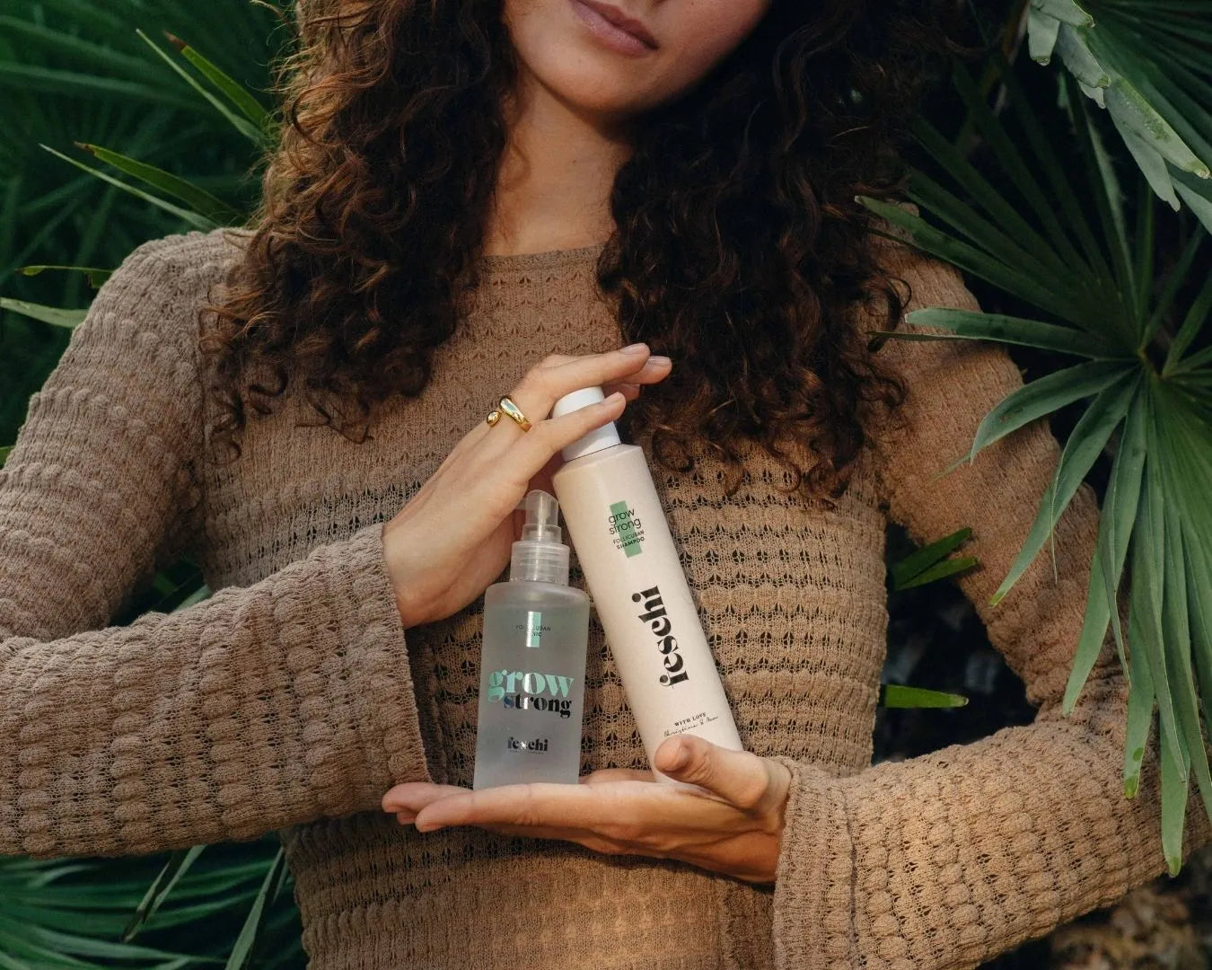Person steht vor grünen Pflanzen mit braunem Kleid und lockigem Haar und hält zwei Produkte von der feschi grow strong - Serie in der Hand