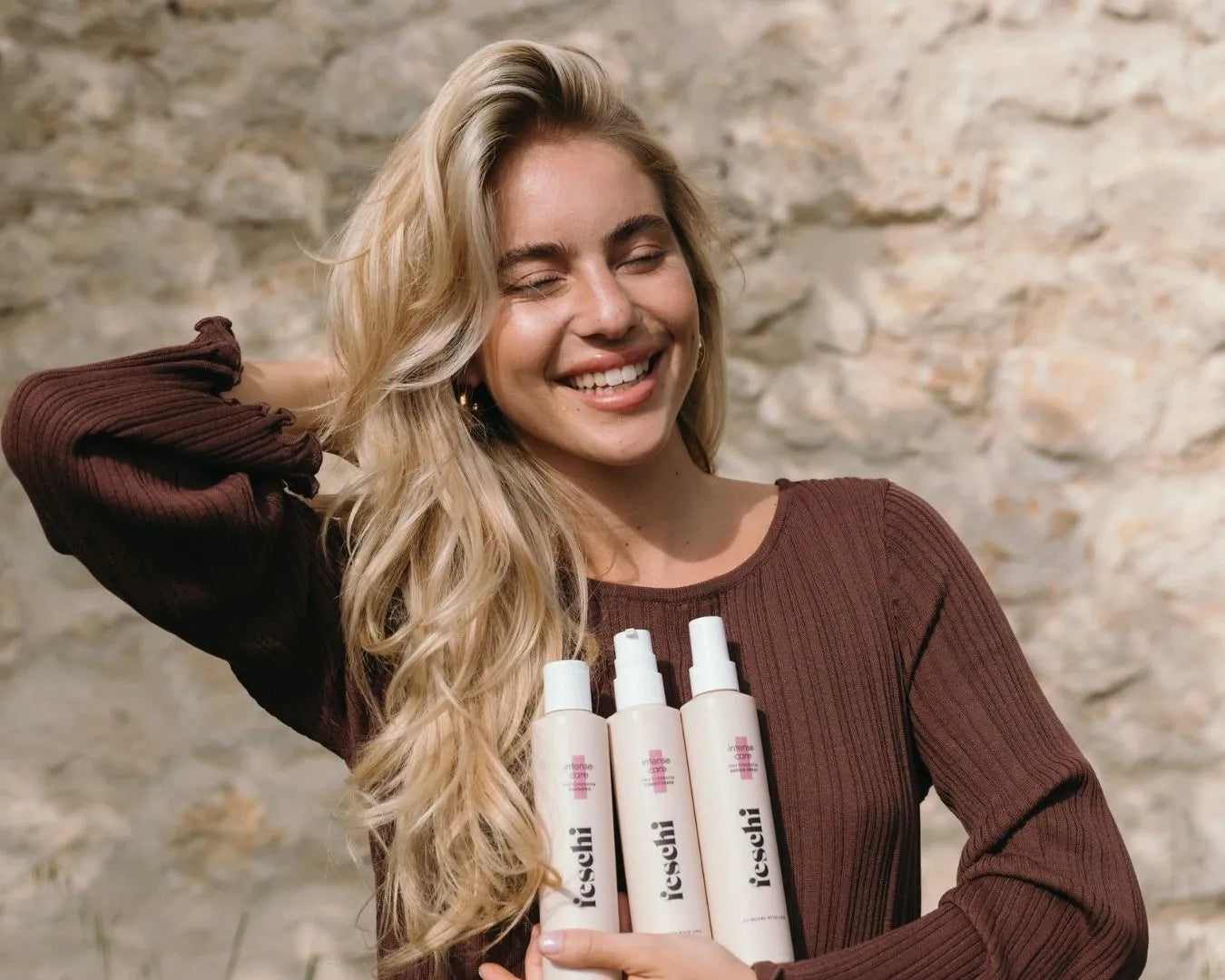 Frau mit blonden, langen Haaren hält drei feschi Produkte von der intense care - Serie in der Hand und steht vor Steinmauer