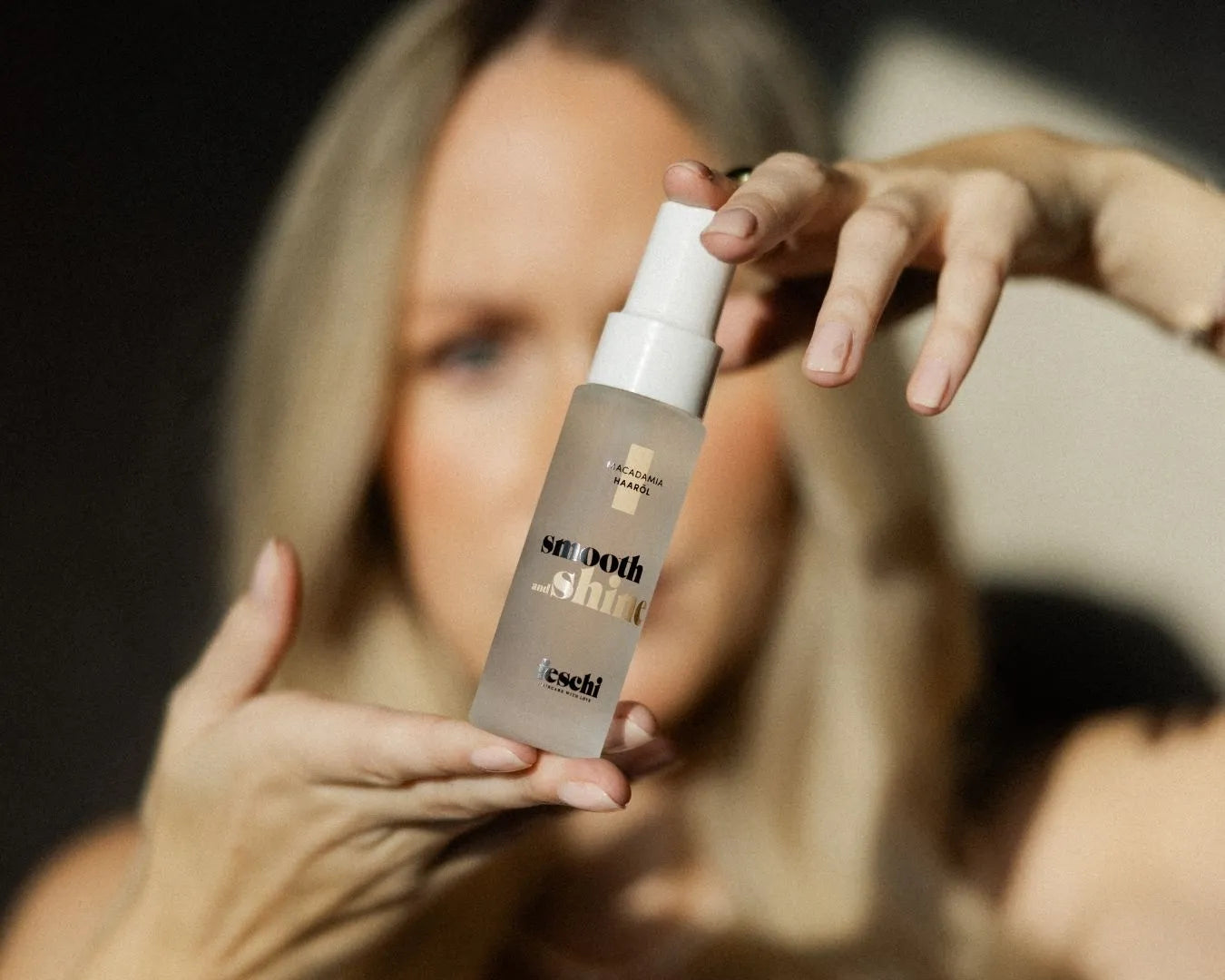 Das feschi Smooth & Shine Haaröl wird von zwei Händen gehalten, im Hintergrund unscharf eine blonde Frau