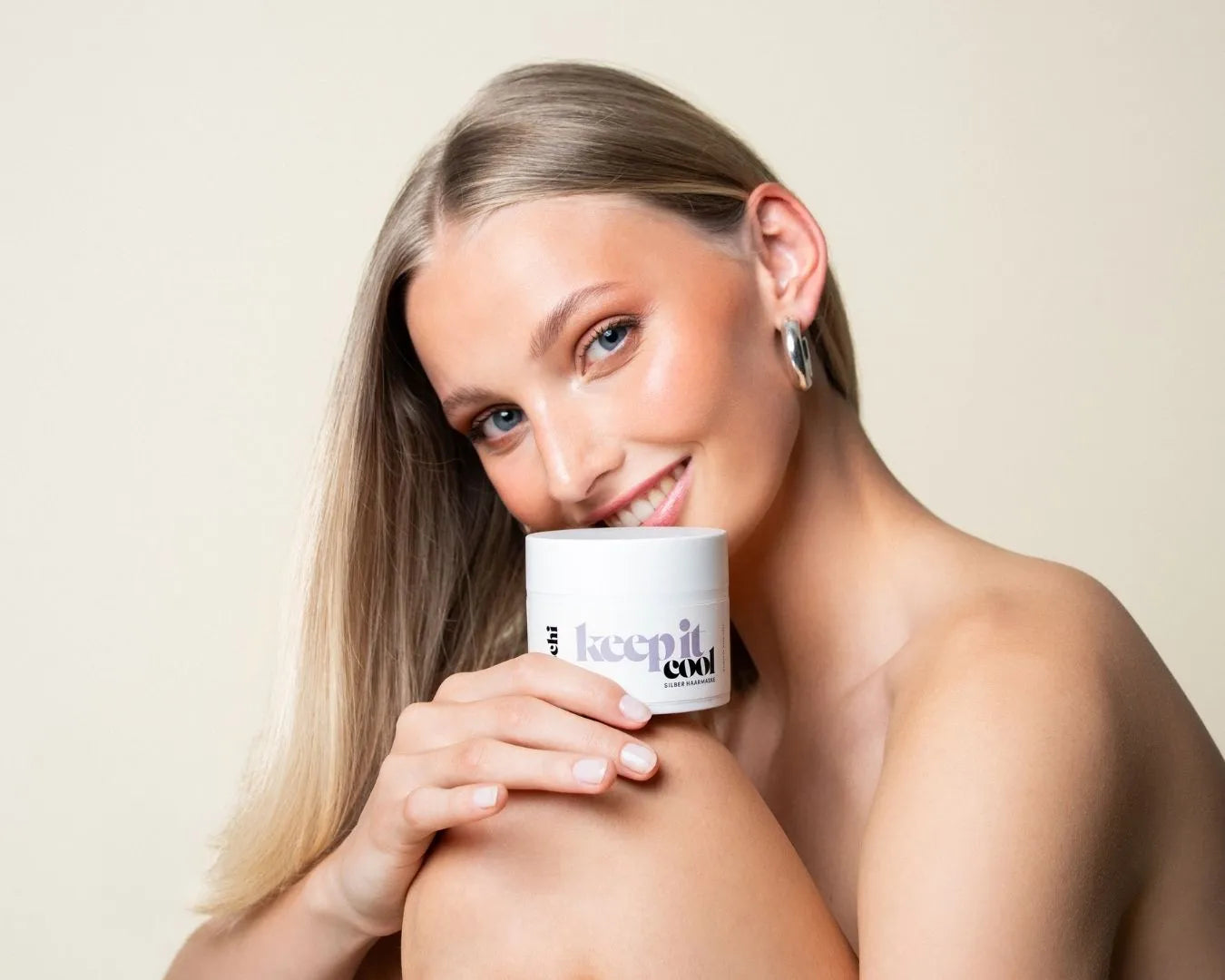 Frau mit langen blonden Haaren, lächelnd in die Kamera, sitzt mit angezogenem Knie, darauf steht die feschi Keep It Cool Silver Mask
