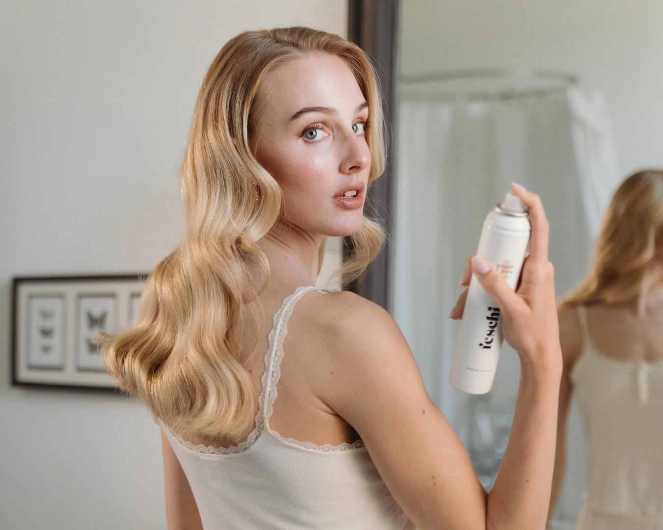 Frau mit blonden, gelockten Haaren hält das Anti Frizz Spray von fesch in der Hand und blickt in Kamera
