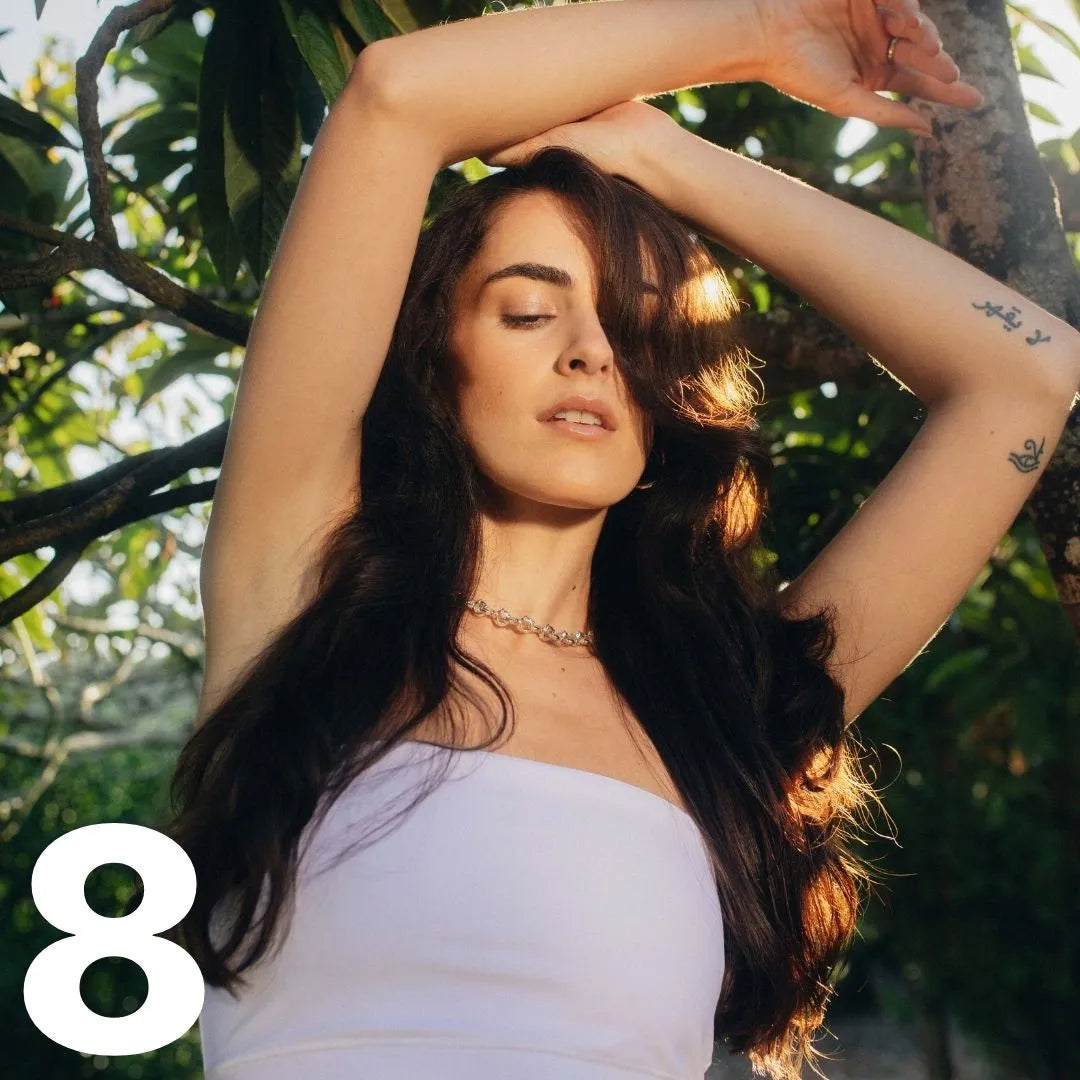 Frau von vorne mit langen braunen Haaren steht im Freien und verschränkt die Arme über dem Kopf