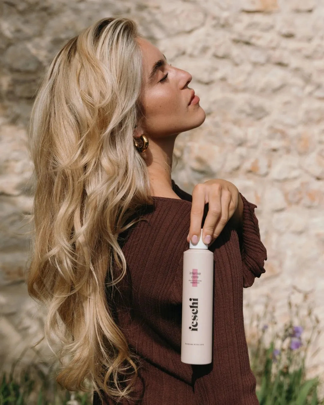 Frau mit langen, blonden Wellen hält das feschi Phytokeratin Shampoo vor natursteinfarbener Wand