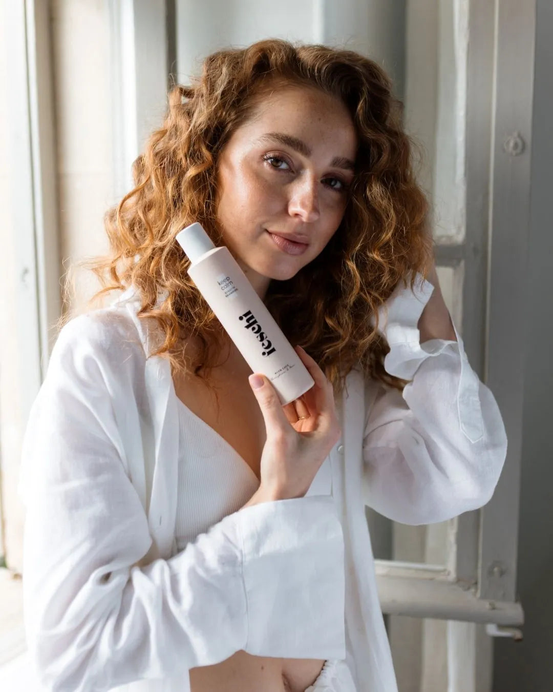Lockige Frau mit natürlichem Look präsentiert das feschi keep calm Sensitiv Shampoo vor einem Fenster