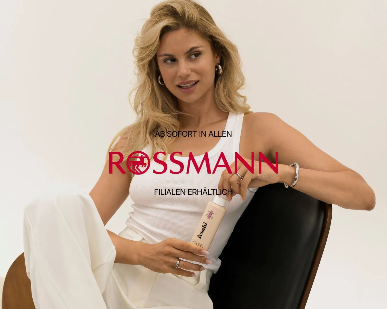 blondes Model sitzt in weißem Outfit auf schwarzem Stuhl und haelt feschi intense care Repair Spray in den Haenden
