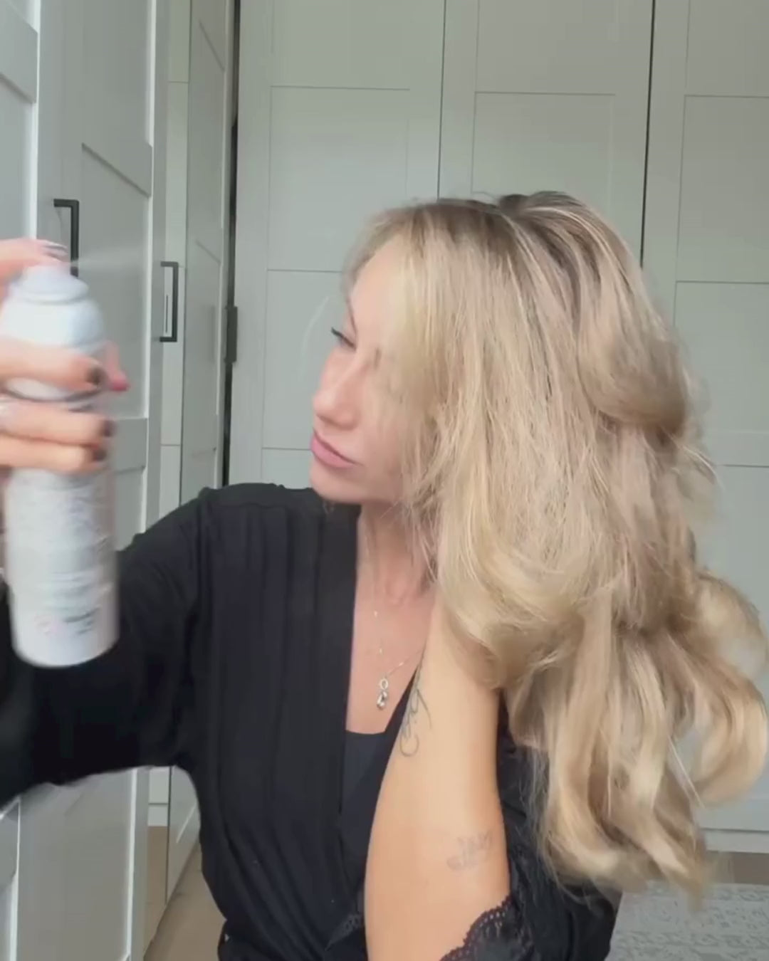 Blonde Frau sprüht sich Haarspray in die Haare und fährt mit Händen durch ihr gestyltes, voluminöses Haar