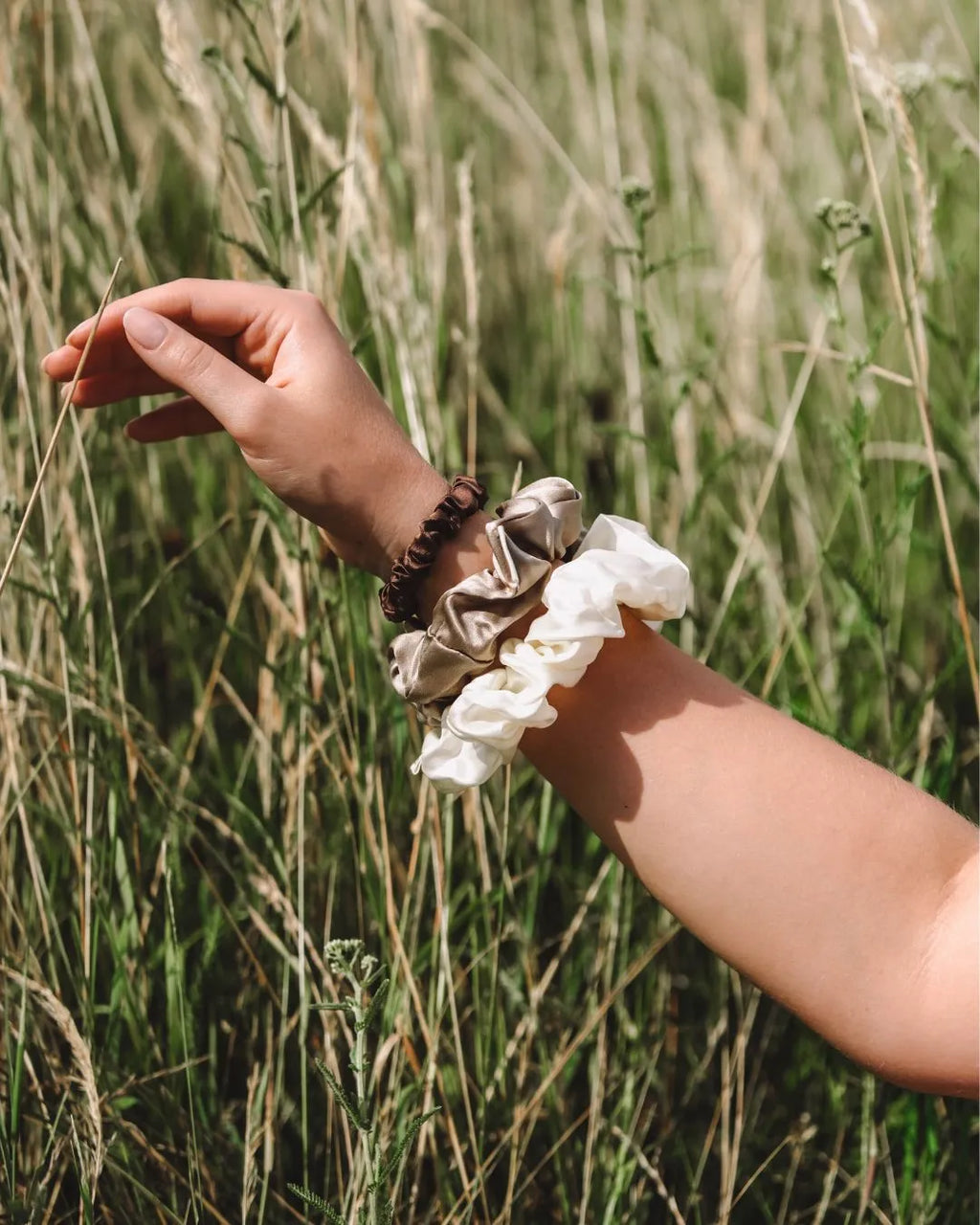 Arm mit dem feschi The Silk Edit Seidenscrunchie Trio, im Hintergrund Wiese