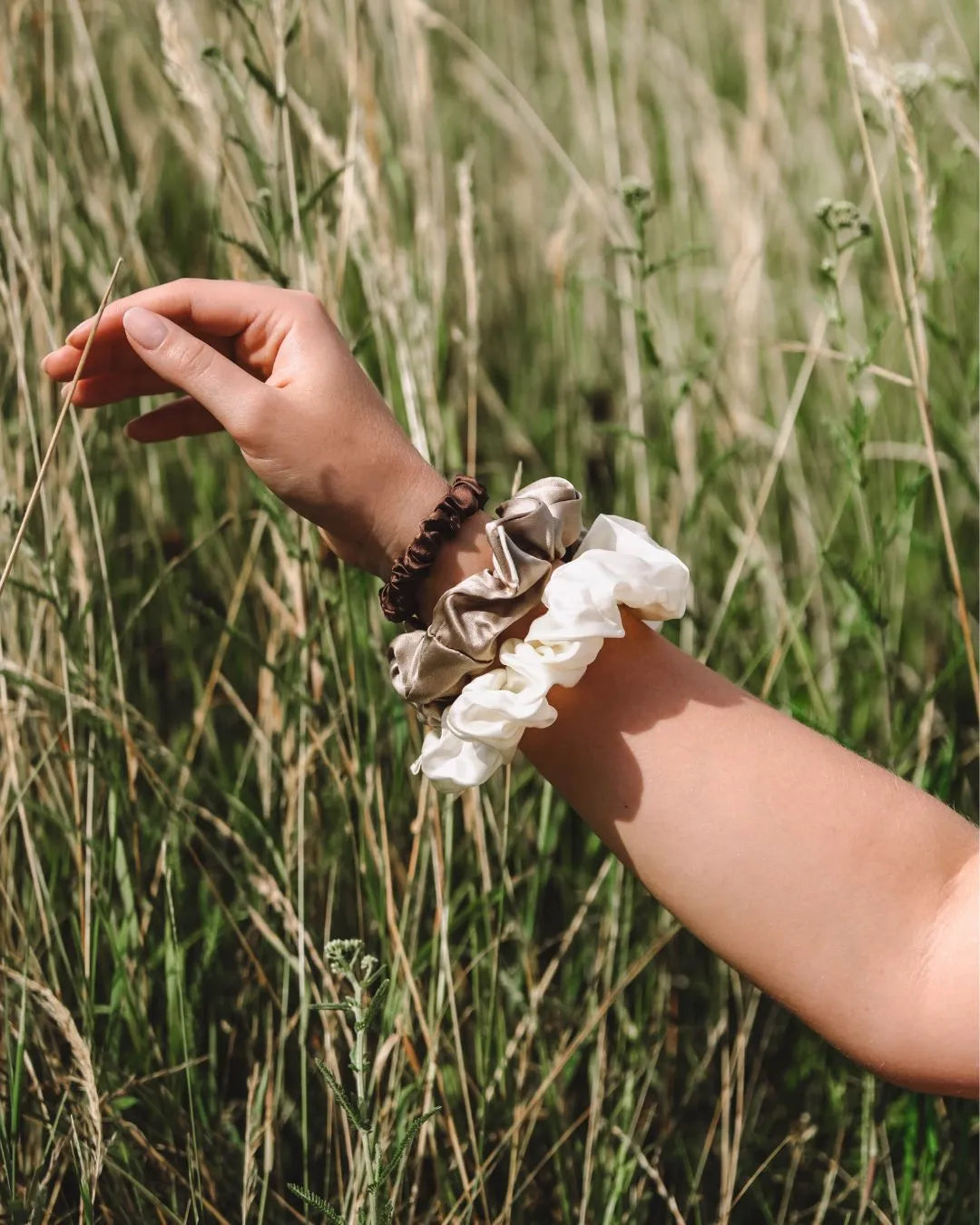Arm mit dem feschi The Silk Edit Seidenscrunchie Trio, im Hintergrund Wiese