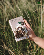 Eine Hand hält das feschi The Silk Edit Seidenscrunchie Trio inklusive Verpackung, im Hintergrund Wiese