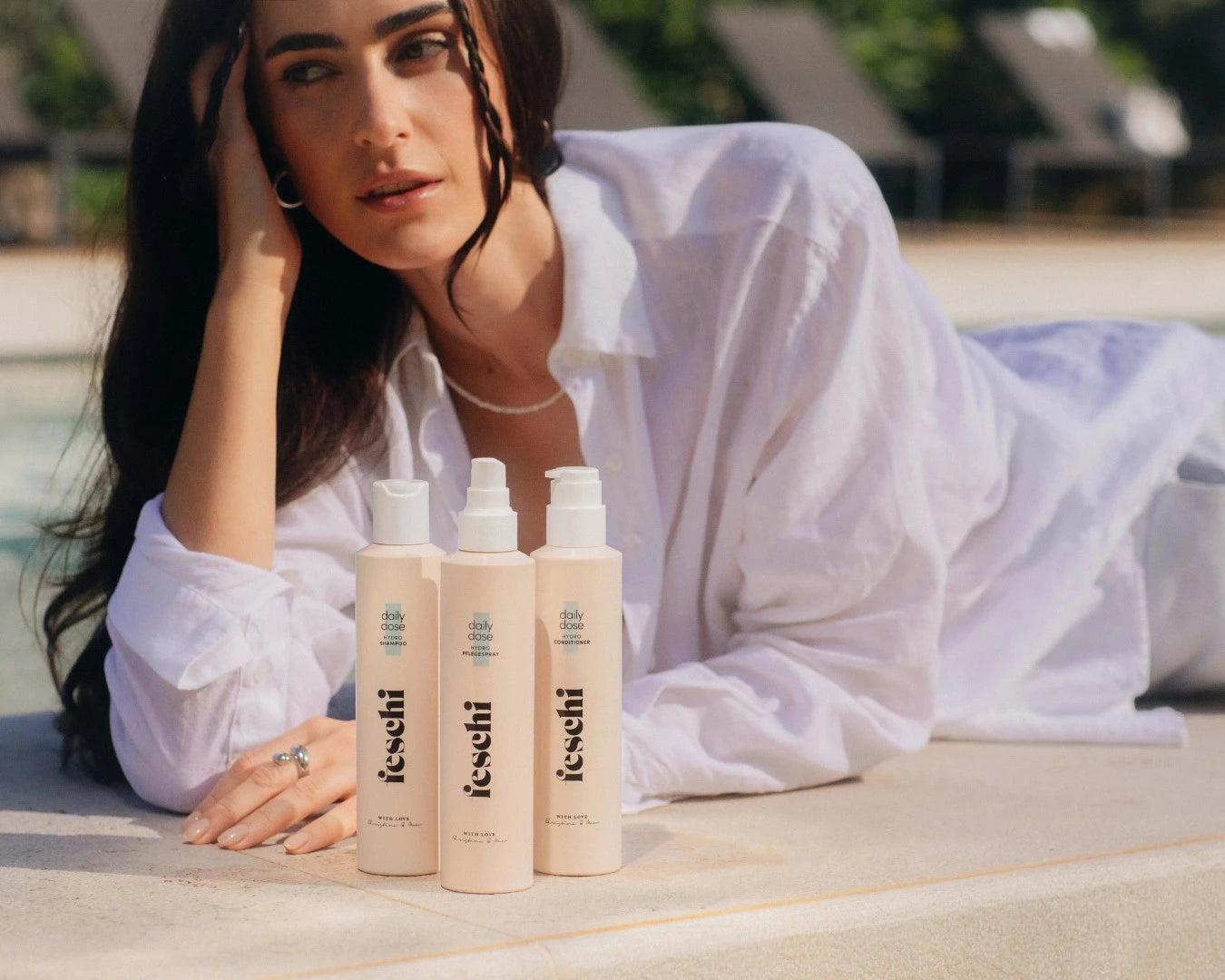dunkelhaarige Frau liegt beim Pool und schaut aus dem Bild raus. Vor ihre stehen das daily dose Shampoo, Conditioner und Pflegespray von feschi. Die Produkte sind im Fokus