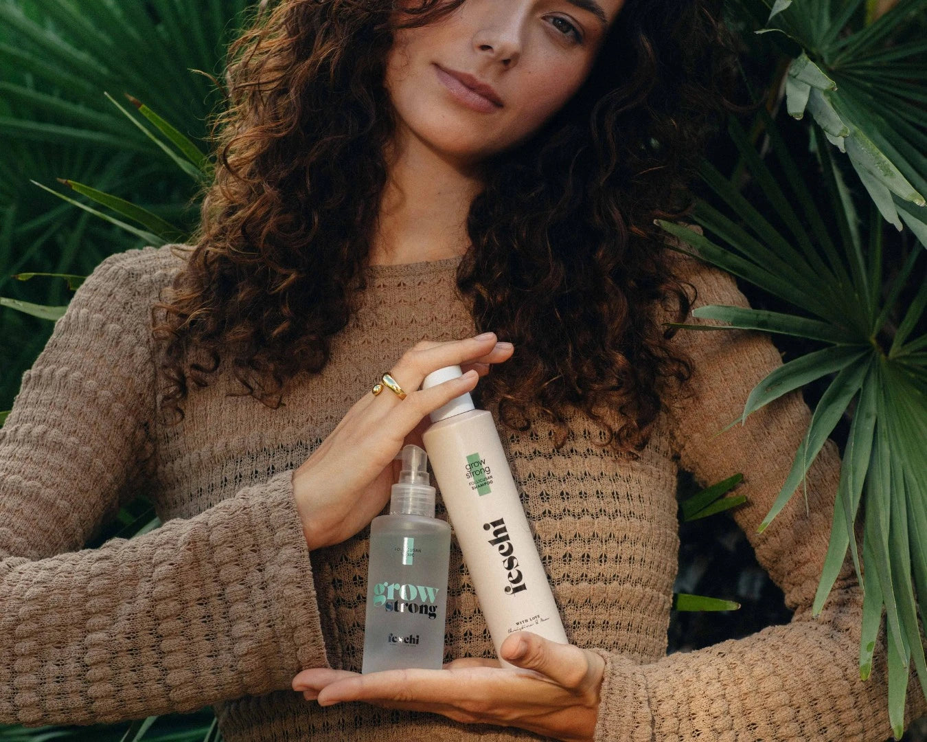 braunhaarige lockige Frau steht vor grüner Pflanze und präsentiert das grow strong Shampoo und Tonic von feschi in ihren Händen