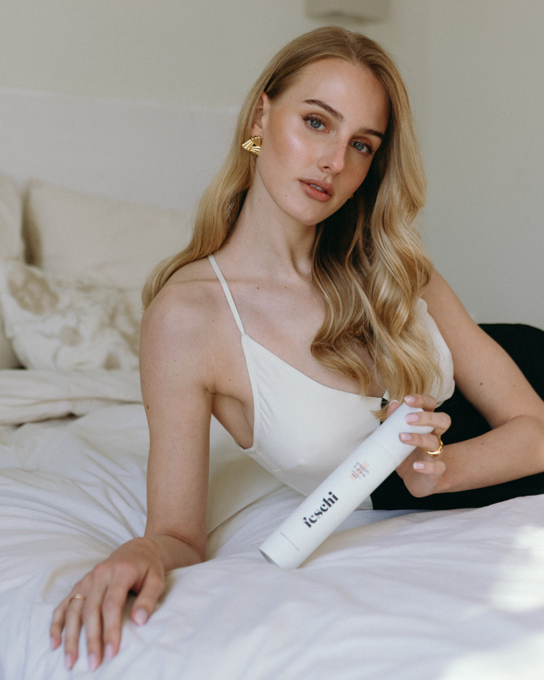 Frau mit blond, gelocktem Haar liegt am Bett und präsentiert den feschi smooth & shine Anti Frizz Spray - für glänzendes Haar und weniger Frizz.
