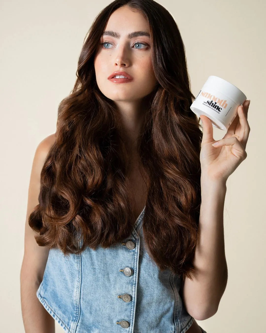 Frau mit langen, welligen & dicken Haaren hält smooth & shine - Macadamia Maske von feschi in der Hand