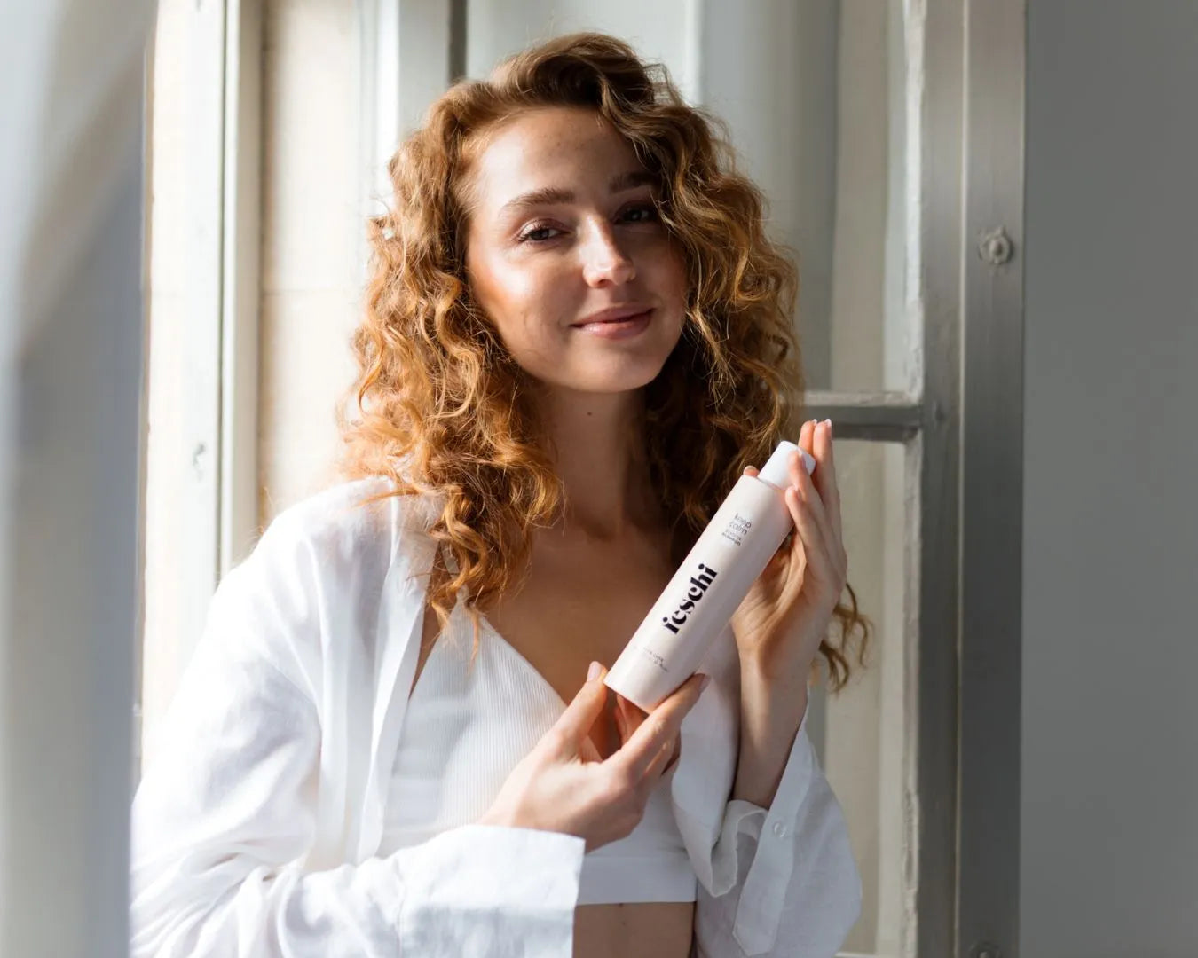 Frau mit Locken, stehend vor einem offenen Fenster, hält das feschi Smooth & Shine Shampoo in der Hand