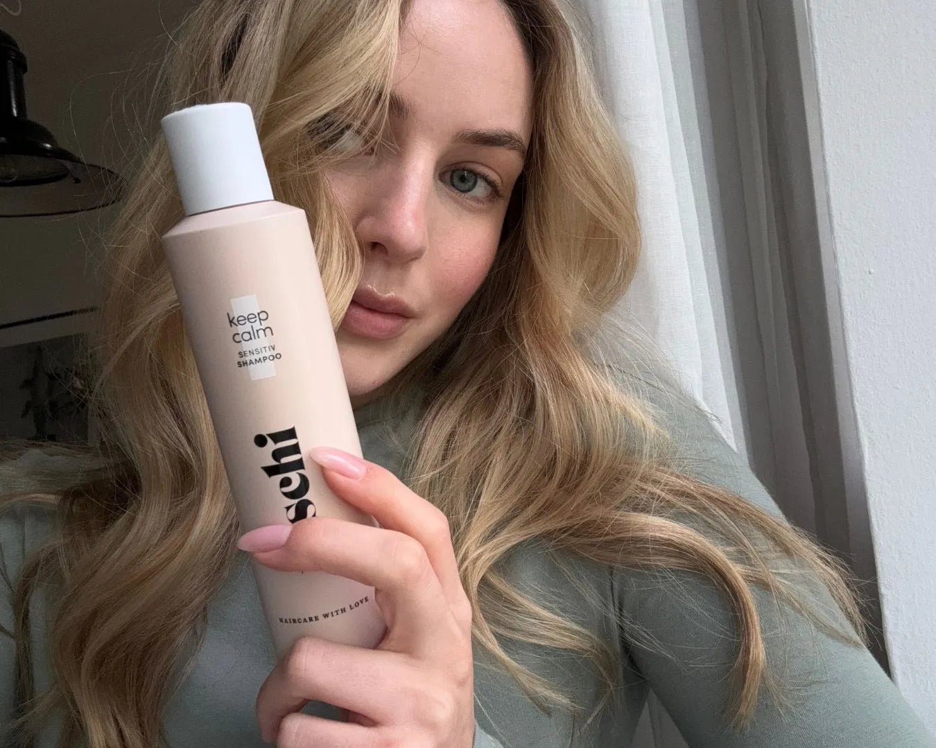 Frau mit blonden Wellen blickt in die Kamera und hält das feschi Keep Calm Sensitiv Shampoo in der Hand