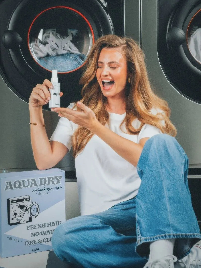 Frau sitzt vor Waschmaschine und hält das aqua-dry Trockenshampoo Liquid von der Marke feschi Haircare in der Hand
