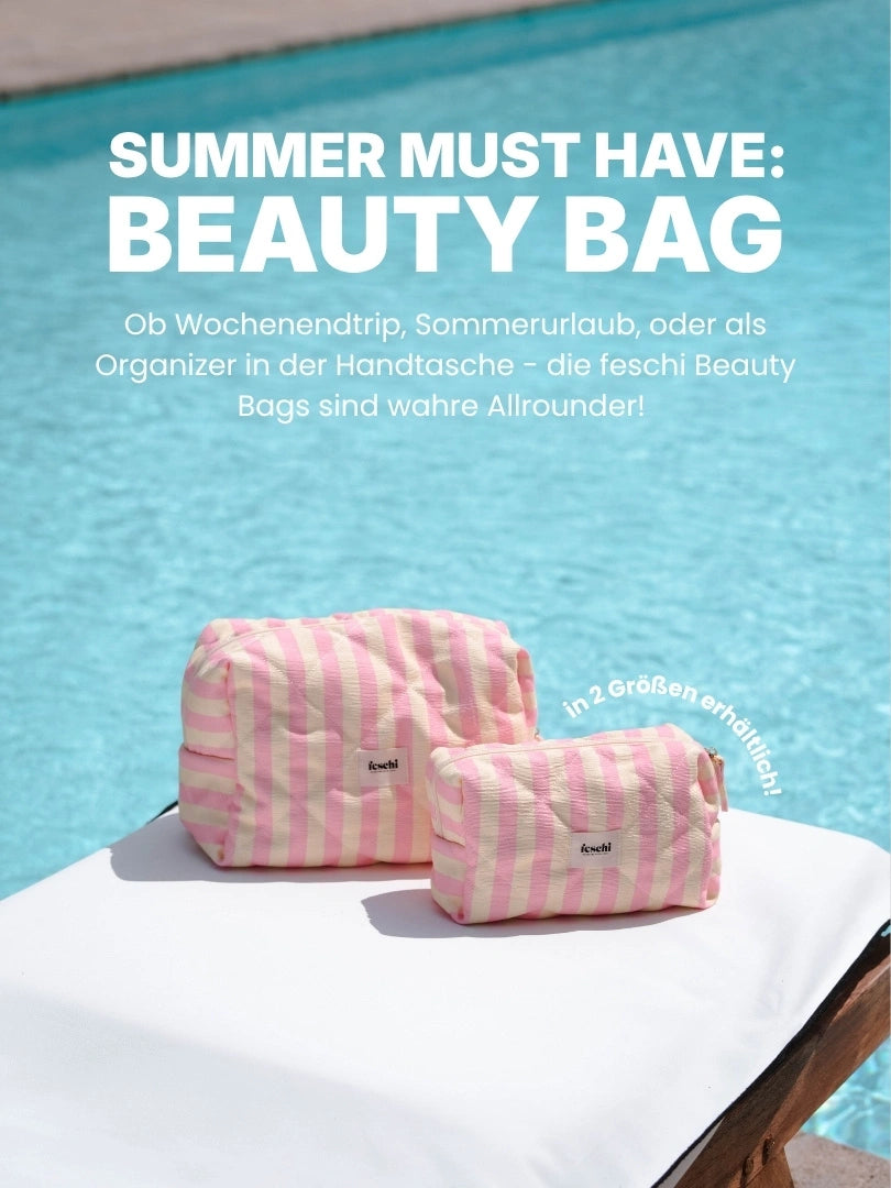 zwei pink gelb gestreifte Kosmetiktaschen von feschi stehen auf Sonnenliege mit weißen Pölstern vor Pool