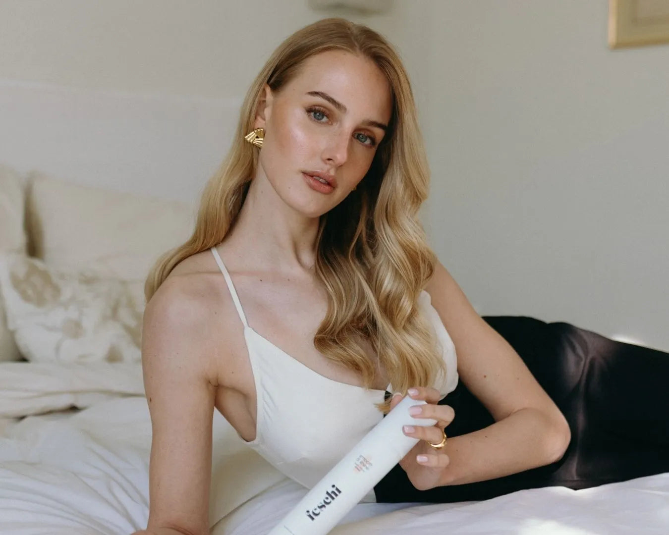 Frau lehnt seitlich im Bett und hat blonde lange Locken und hält das Anti Frizz Spray in der Hand